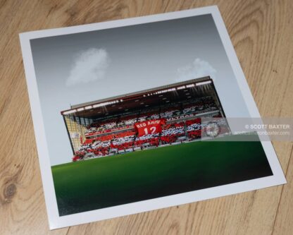 A photograph of Aberdeen Football Club - Pittodrie Stadium by Aberdeen photographer Scott Cameron Baxter. The image is taken from the merkland road end, towards the Richard Donald Stand. The brass is green and the stand is full with supporters holding a RED ARMY banner shaped like a t-shirt. it is red. the fans have mixed red and white paper qurares all aroudn the stand. There are two clouds above the stand and the sky is gray.